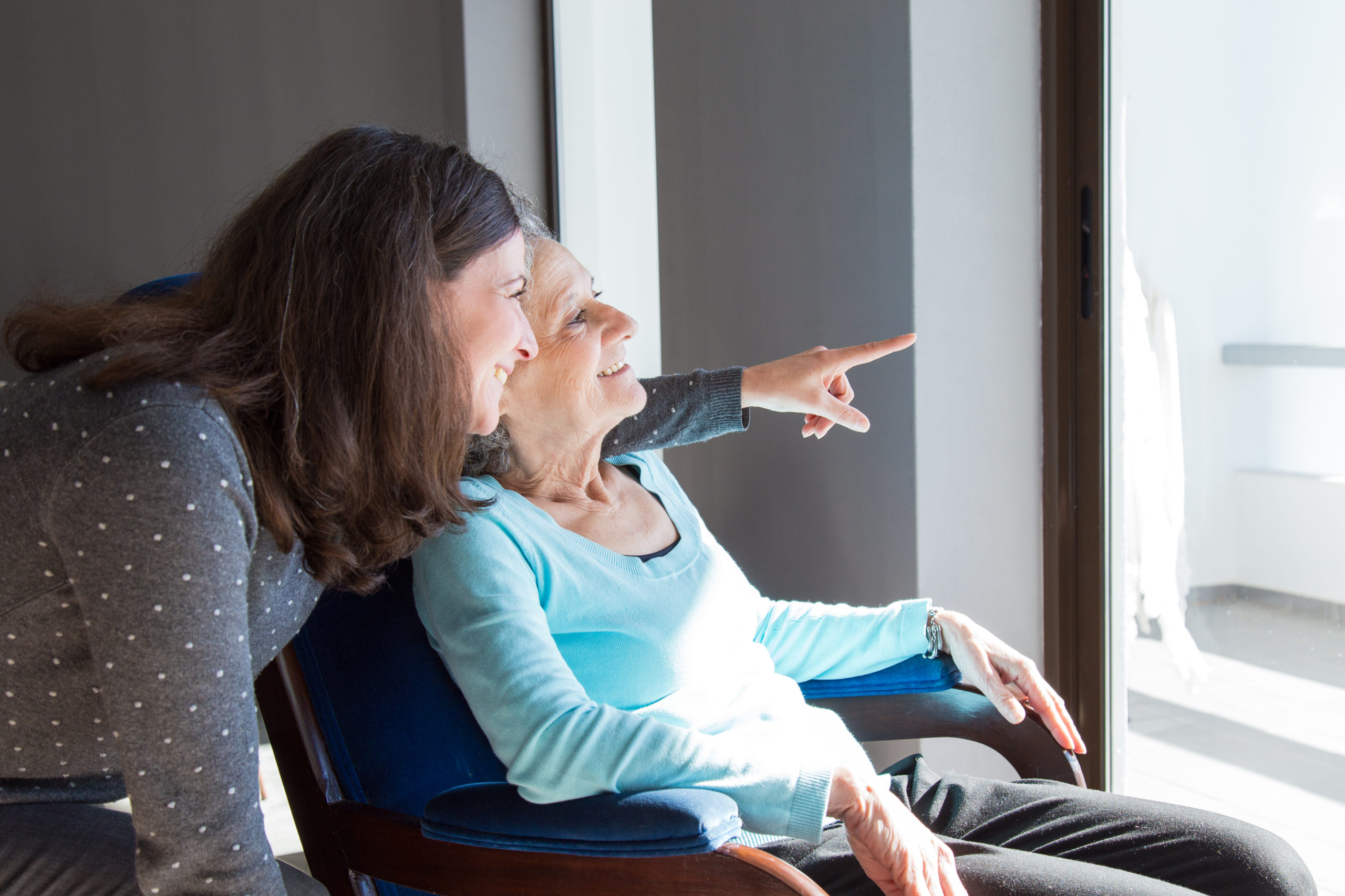 Positive mother and daughter enjoying dramatic view out of window. Young health visitor showing to senior lady funny scene out of window. Family relations concept
